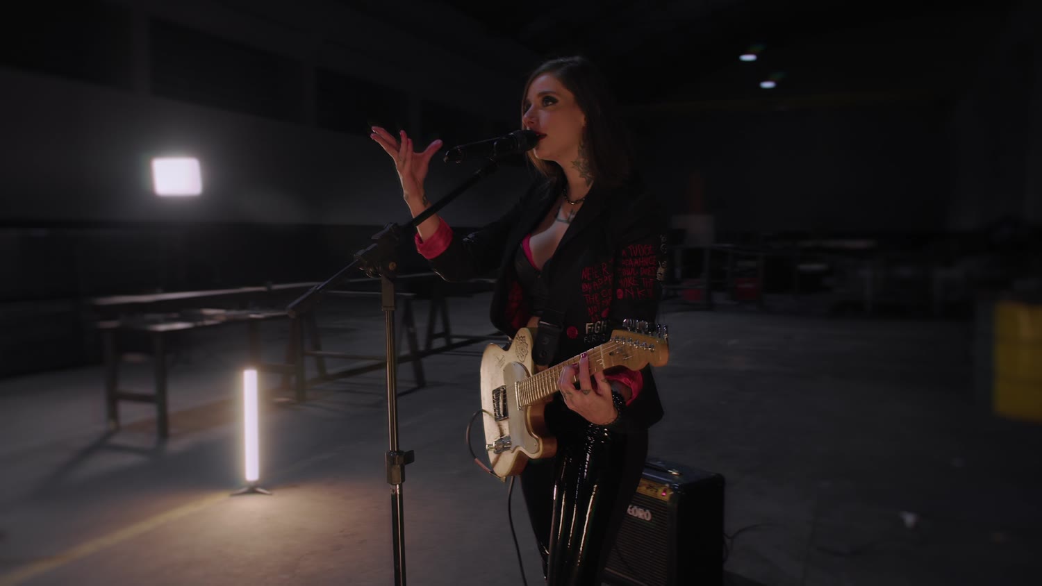 A cantora da Johaine no microfone, com a guitarra, no galpão escuro